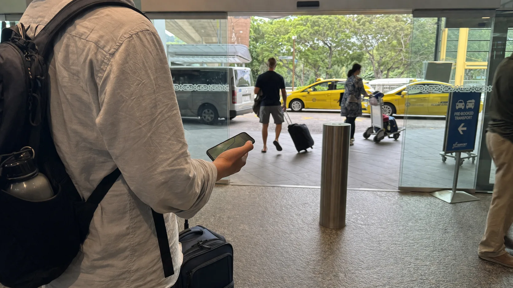 Traveler in an airport terminal holding a smartphone and small suitcase, looking outside at taxis and vans.